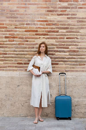 full length of redhead woman in dress holding purse and standing with suitcase near brick wall on european streetの写真素材