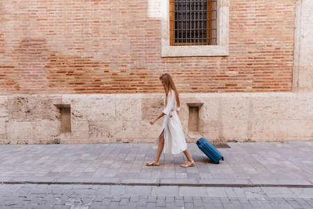 full length of redhead woman in dress walking with suitcase near brick wall on european streetの写真素材