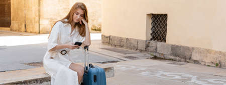 redhead woman in dress sitting near suitcase and using cellphone in valencia, bannerの写真素材