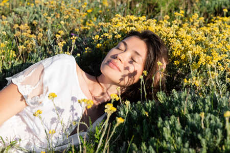 Brunette woman in white dress lying near flowers and grass on meadowの写真素材