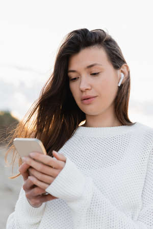 Brunette woman in wireless earphone using blurred smartphone on beachの写真素材