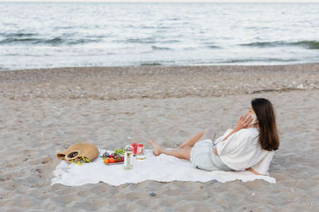 Young woman talking on smartphone near wine and fruits on blanket on beachの写真素材