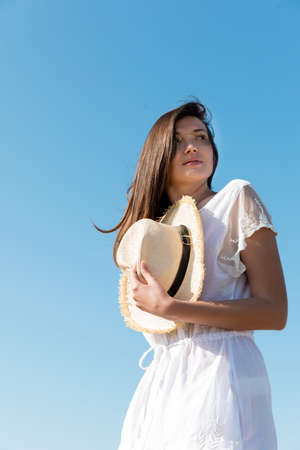 Low angle view of young woman in dress holding sun hat and looking away with sky at backgroundの写真素材