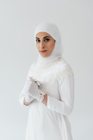 portrait of muslim woman in white hijab and wedding dress looking at camera isolated on greyの写真素材