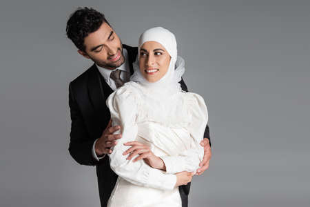 smiling groom standing behind cheerful bride in wedding dress isolated on greyの写真素材