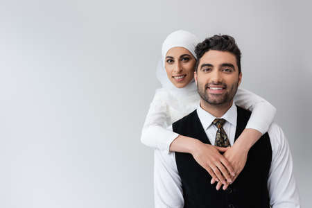 happy muslim bride in hijab hugging cheerful groom in suit isolated on greyの写真素材