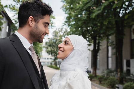 happy muslim bride in white hijab and groom in suit looking at each otherの写真素材