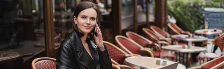 smiling woman in black jacket talking on smartphone in french outdoor cafe, bannerの写真素材