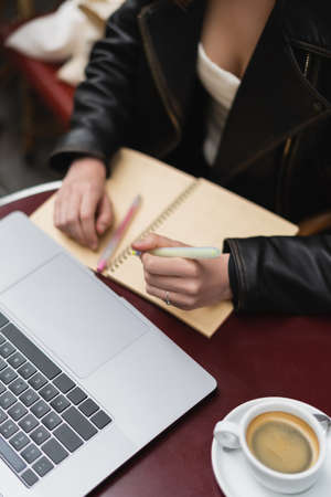 cropped view of freelancer making notes near laptop and cup of coffee on table in french outdoor...
