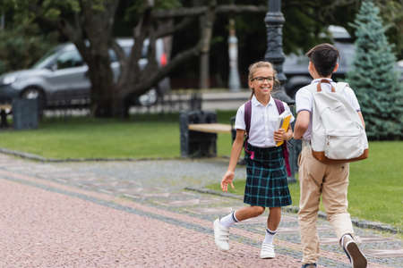 Positive schoolkid in eyeglasses talking to friend with backpack outdoorsの写真素材