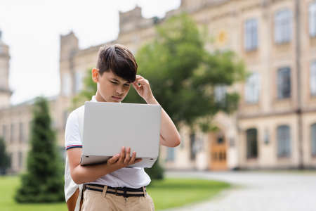 Pensive asian schoolboy looking at laptop outdoorsの写真素材