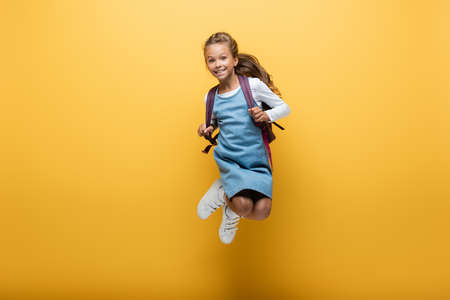 Happy preteen schoolgirl with backpack jumping on yellow background