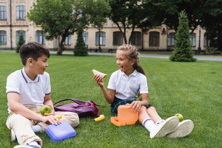 Excited schoolgirl holding sandwich near lunchbox and asian friend with apple on lawnの写真素材