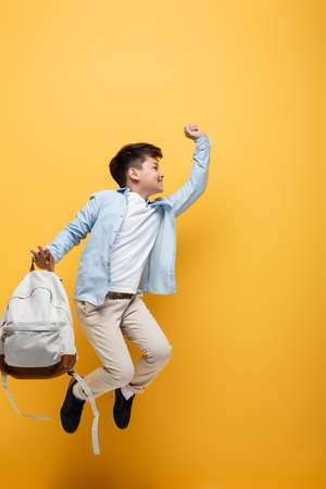 Side view of asian schoolboy holding backpack and jumping isolated on yellow