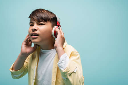 Focused asian schoolchild listening music in headphones isolated on blue