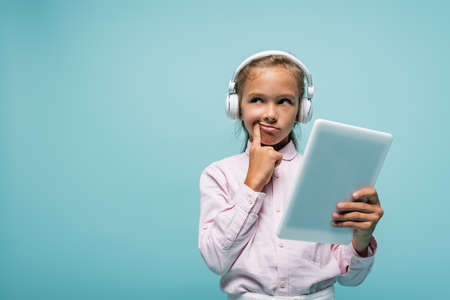 Pensive schoolkid in headphones holding digital tablet isolated on blue