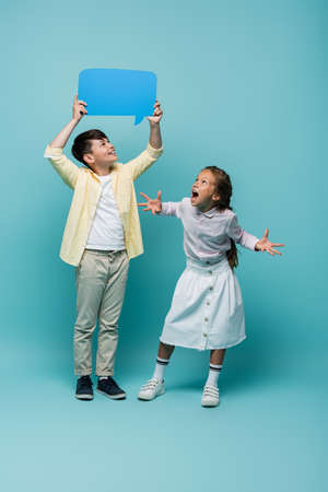 Excited and smiling interracial pupils looking at speech bubble on blue background