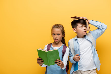 Schoolgirl reading book and sticking out tongue near asian friend isolated on yellowの写真素材