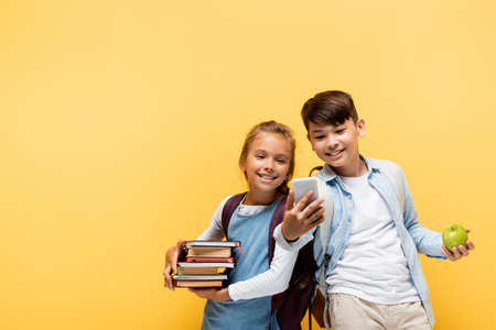 Smiling multiethnic pupils with books and apple using mobile phone isolated on yellowの写真素材