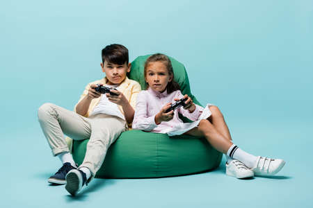 KYIV, UKRAINE - JULY 2, 2021: Focused interracial kids playing video game on beanbag chair on blue...