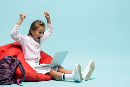Excited pupil showing yes gesture while looking at laptop near backpack on blue background