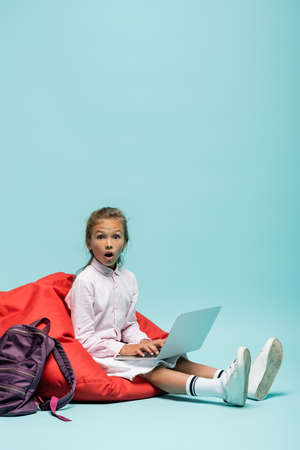Shocked schoolgirl using laptop on beanbag chair on blue background