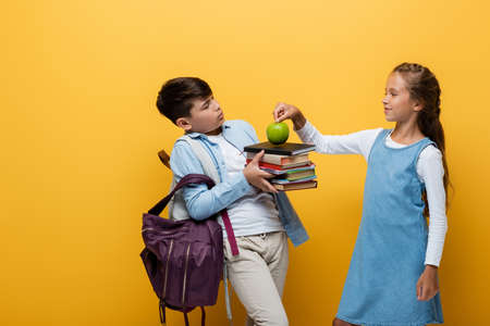 Schoolgirl putting apple on books near asian friend on yellow backgroundの写真素材