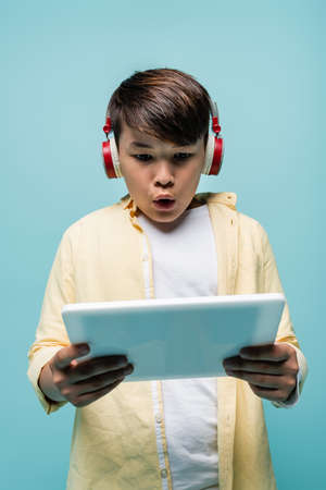 Shocked asian schoolkid in headphones using digital tablet isolated on blueの写真素材