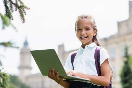 Happy schoolkid holding laptop and looking at camera outdoors