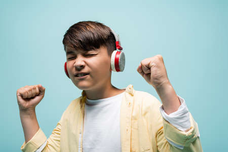 Excited asian child listening music in headphones and showing yes gesture isolated on blue