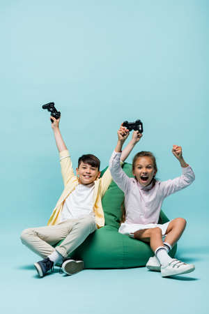 KYIV, UKRAINE - JULY 2, 2021: Excited multiethnic kids holding joysticks on beanbag chair on blue...
