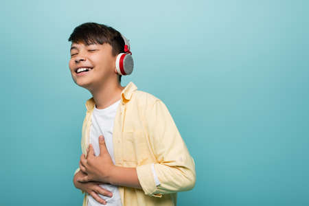 Happy asian schoolkid in headphones smiling isolated on blue
