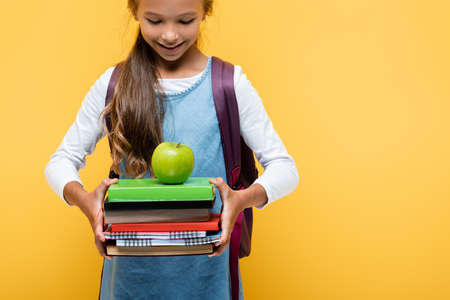Smiling schoolchild looking at books and apple isolated on yellowの写真素材