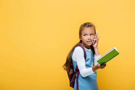 Schoolgirl talking on smartphone and holding book isolated on yellow
