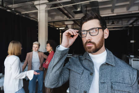 bearded businessman adjusting eyeglasses and looking away while multicultural team talking on blurred backgroundの写真素材