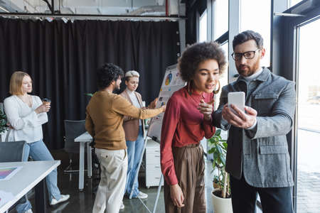 businessman in eyeglasses showing smartphone to african american colleague in advertising agencyの写真素材