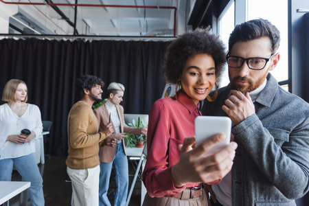 smiling african american woman showing smartphone to thoughtful colleague in ad agencyの写真素材
