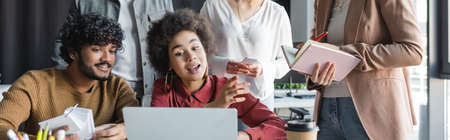 african american woman talking near laptop and indian colleague in advertising agency, bannerの写真素材