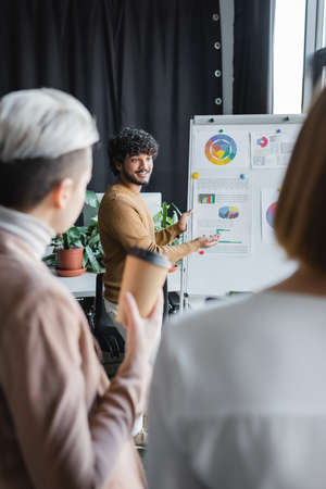 happy indian advertising designer talking to blurred colleagues near flip chart with graphsの写真素材