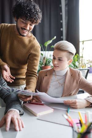 positive indian advertising designer pointing at color samples near colleague in office