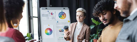 woman with coffee to go talking to blurred interracial colleagues near flip chart in ad agency, bannerの写真素材