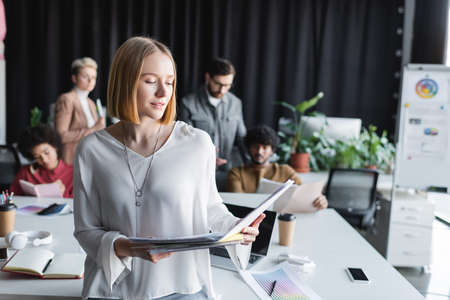 smiling woman looking in folder near blurred interracial team working in advertising agencyの写真素材