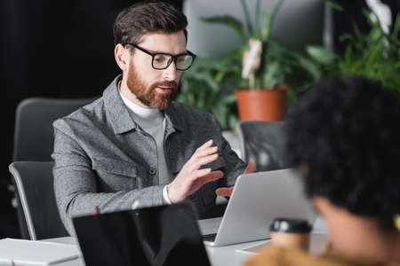 bearded man gesturing near laptop during video call in advertising agency
