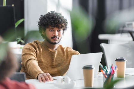 young indian designer working near laptop in advertising agency on blurred foreground