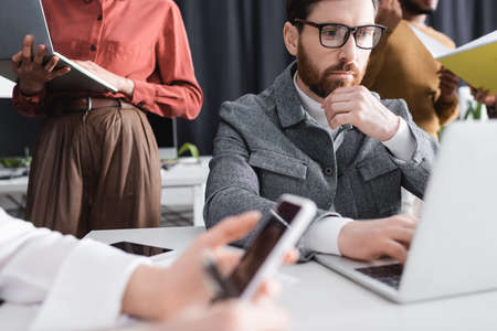 pensive bearded man in eyeglasses working near laptop and blurred multicultural team in ad agency