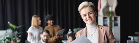 cheerful designer looking at camera near blurred multicultural colleagues in ad agency, banner