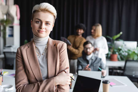 cheerful manager with pen looking at camera near blurred interracial team in ad agencyの写真素材