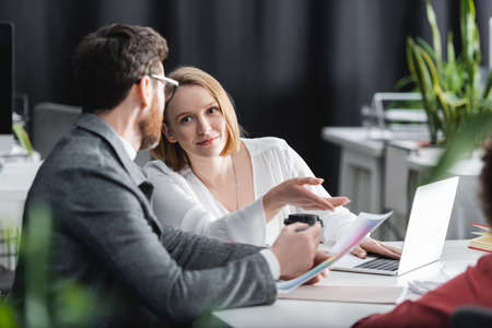 smiling woman pointing with finger near laptop and colleague in ad agency