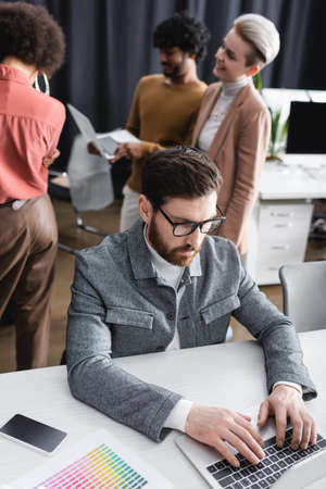 businessman in eyeglasses typing on laptop near multiethnic advertising agents on blurred backgroundの写真素材