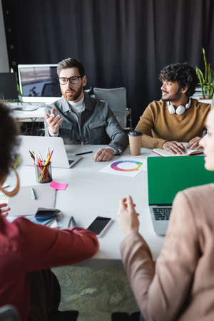 man in eyeglasses pointing at blurred colleagues near smiling indian man in advertising agency